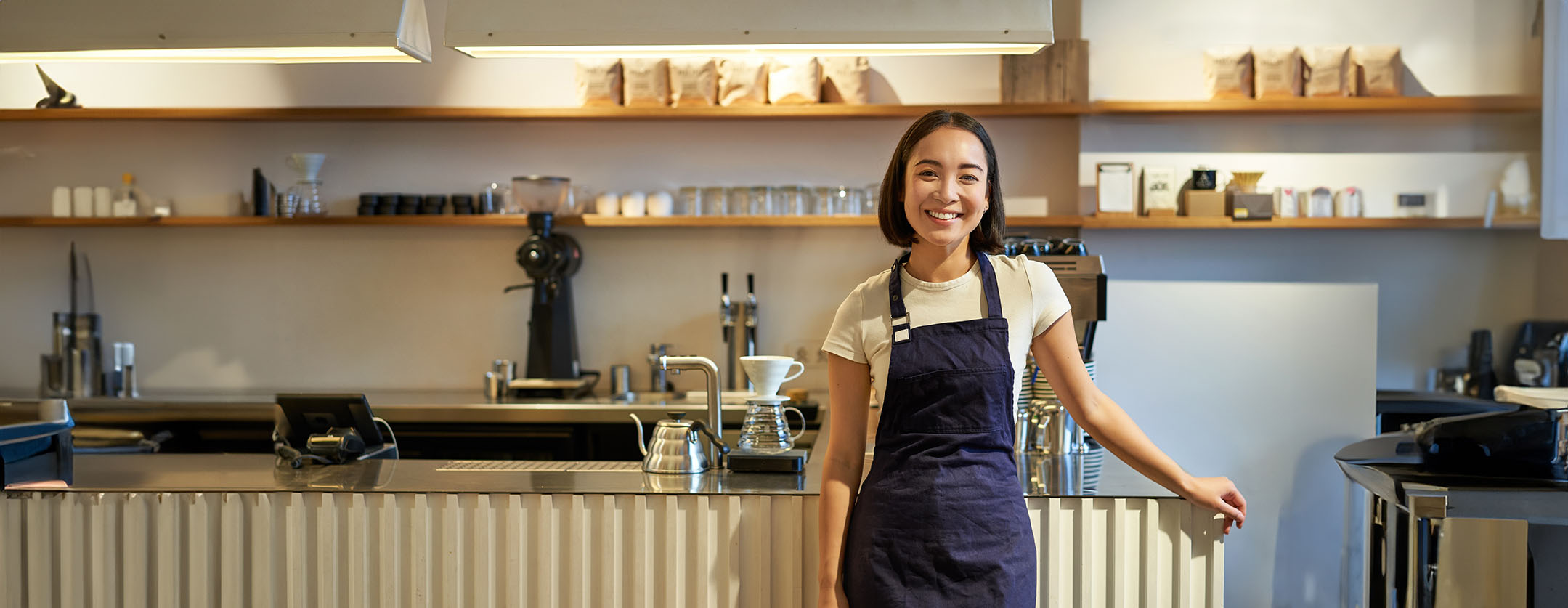 Portrait of smiling asian female barista, wearing apron, standing near counter with coffee, working in cafe