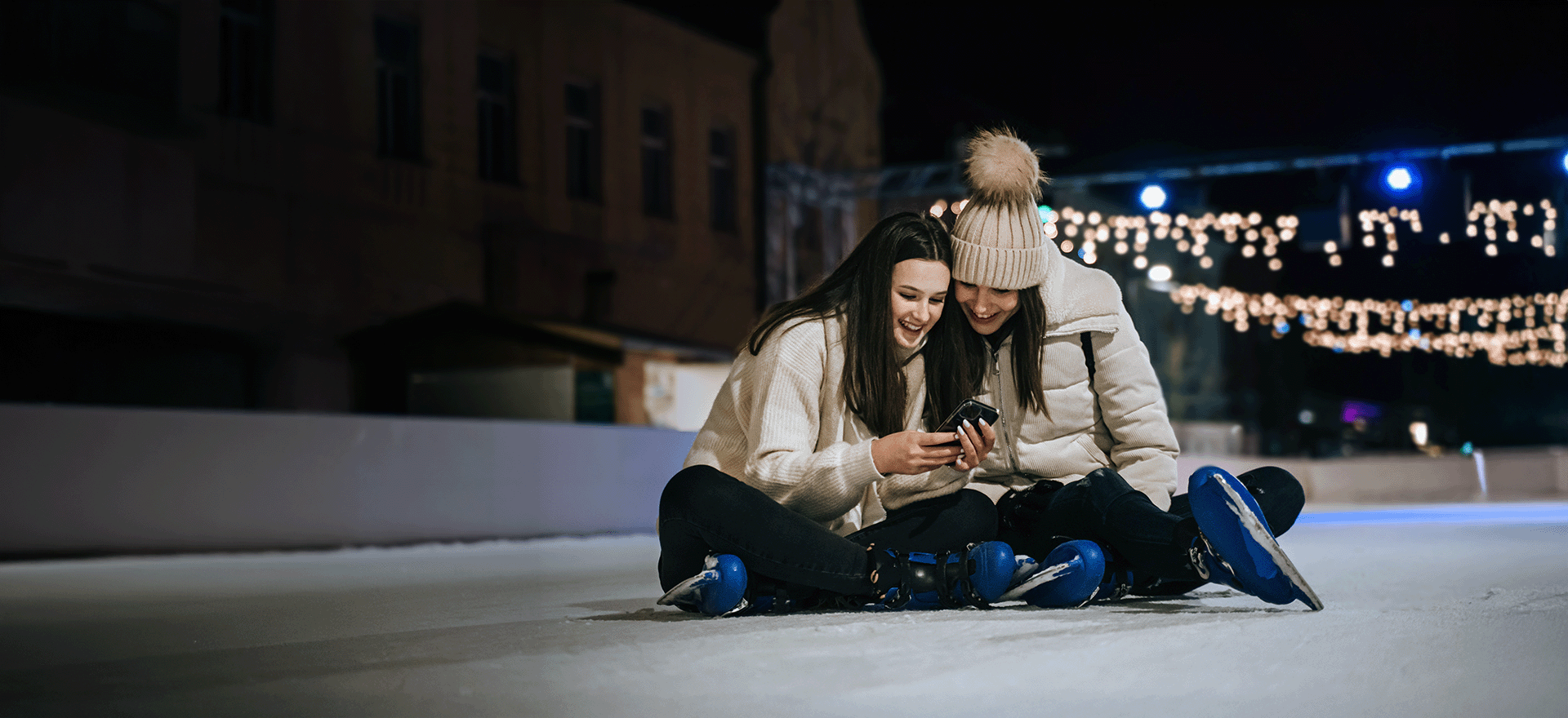 two girls sitting cross legged on an ice rink looking and their phones together